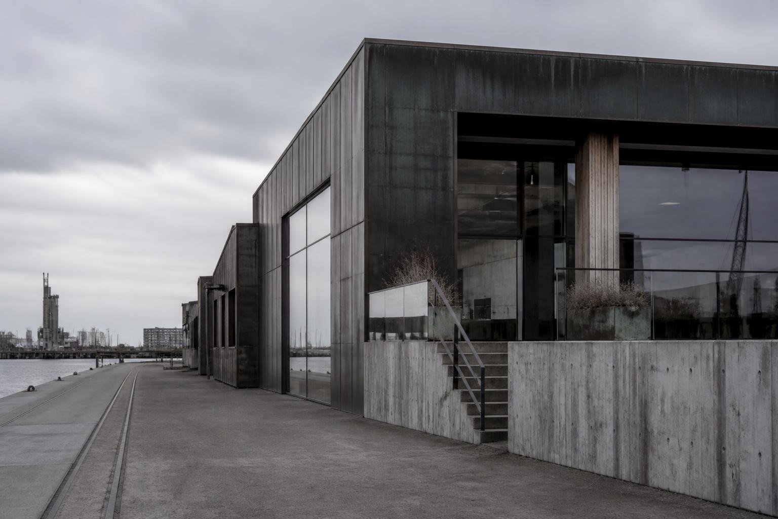 Ombygget pakhus i rødbrun tegl med et regelmæssigt vinduesgitter over en mørk, glasindrammet stueetage ud mod havnen.