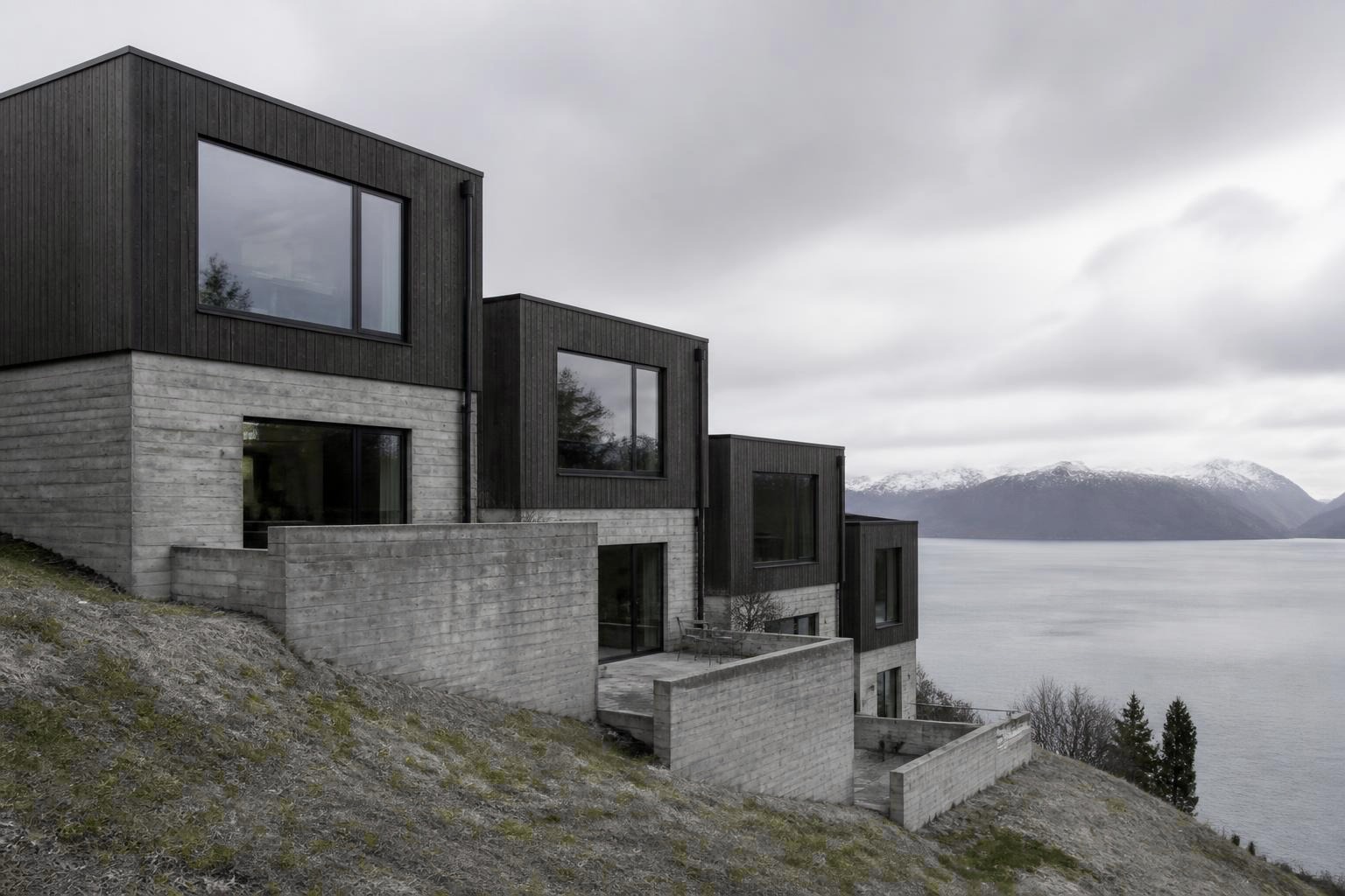 Terrasseret hus i beton og træ på en grøn skrænt ned mod en fjord, i køligt dagslys.