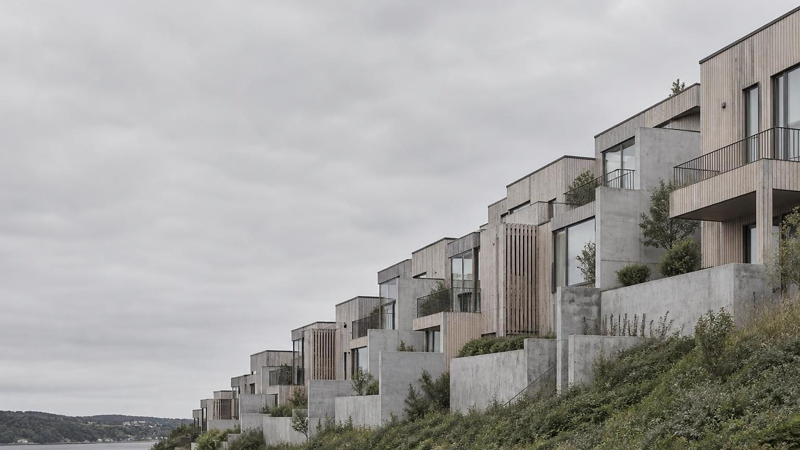 Terrasseret boligbebyggelse i beton og træ, opført i et let skrånende landskab under overskyet himmel.